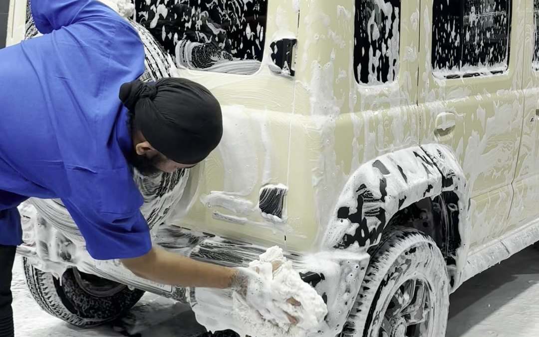 A team member hand washing a foam-covered Suzuki Jimny with a wash mitt